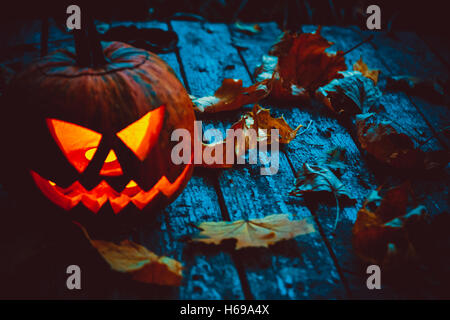 Leuchtender Kürbis, der den Kopf des alten Jack symbolisiert, mit Herbstlaub auf hölzernem Hintergrund. Weichzeichner. Oberer Freiheitsgrad Stockfoto