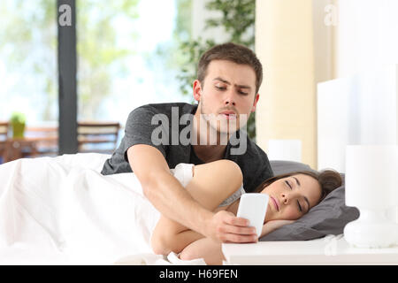 Eifersüchtiger Ehemann Spionage Handy von seinem Partner, während sie in einem Bett zu Hause schläft Stockfoto