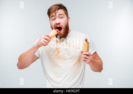 Hungrigen bärtiger Mann in schmutziges Hemd beißen zwei Hotdogs isoliert auf weißem Hintergrund Stockfoto
