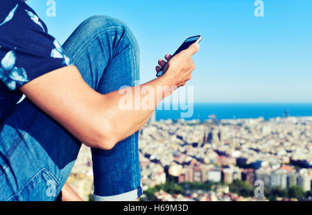 Closeup von einem kaukasischen Jüngling mit seinem Smartphone in der Spitze eines Hügels in Barcelona, Spanien, mit Panoramablick auf das cit Stockfoto