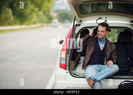 Freunde anzurufen. Glücklich Jüngling Handy sprechen und sitzt auf einem Kofferraum eines Autos Stockfoto