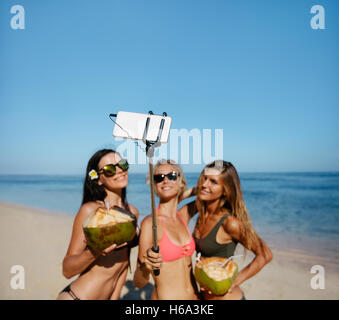 Drei junge Frauen im Bikini am Strand unter Selbstbildnis mit Smartphone auf Selfie Stick. Gruppe von Freundinnen halten Stockfoto