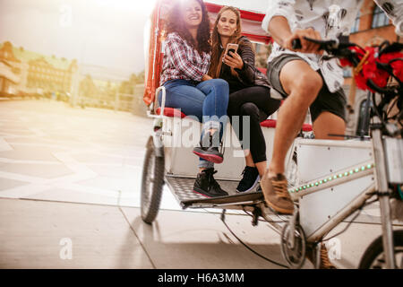 Junge Frauen sitzen auf Dreirad und posieren für Selfie. Freundinnen genießen Dreirad fahren auf der Straße und nehmen Selbstporträt wi Stockfoto