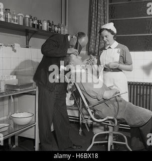 1950, historische, eine männliche Arbeiter in seinem Overall sitzt auf einem Stuhl in der Chirurgie des Unternehmens oder medizinische Zimmer seine Augen von einem männlichen Arzt geprüft, mit einem uniformierten Krankenpflege Schwester in Support, England, UK. Stockfoto