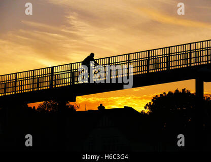 Person in der Silhouette, Radfahren über Fußgänger Fußgängerbrücke mit Farbe gefüllt Sonnenuntergang Himmel dahinter. Stockfoto