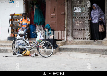 Ein Mann flickt ein Fahrrad vor einem Geschäft, während Frauen in ein Kopftuch und ein Khimar in Stonetown Sansibar beobachten Stockfoto