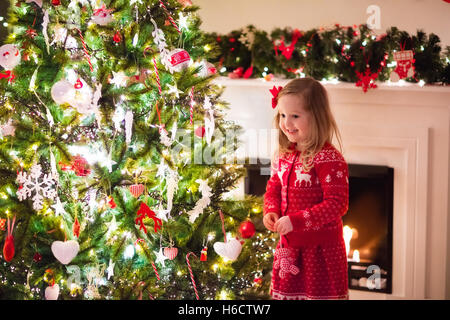 Kleines Mädchen in rot Strickpullover nordische Rentier hängende Ornamente auf Weihnachtsbaum mit Licht, Tand und Candy Canes. Stockfoto