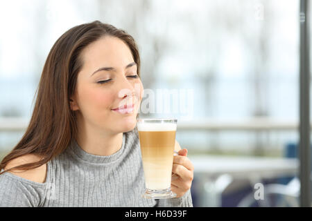 Frau genießt riechen und halten einen Macchiato Kaffee in einem Restaurant mit einem Fenster mit dem Meer im Freien im Hintergrund Stockfoto