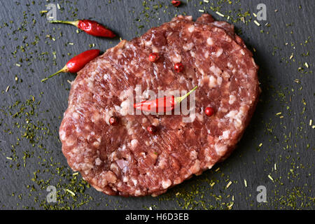 Nahaufnahme von rohen Burger gewürzt mit roten Pfefferkörner, Chilischoten und Kräutern auf eine Schieferoberfläche Stockfoto