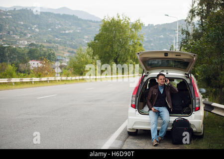 Freunde anzurufen. Glücklich Jüngling Handy sprechen und sitzt auf einem Kofferraum eines Autos Stockfoto