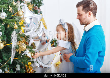 Tochter und Vater schmücken Christbaum. Vater Tochter unterstützt und kleine süße Mädchen hängt Kugeln auf dem Weihnachtsbaum. Stockfoto