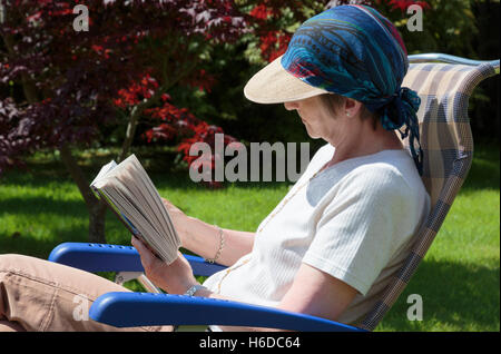 Jemand pensionierte ältere Frau entspannen in einem Garten im Hinterhof Stuhl ein Buch lesen im Sommer Sonnenschein im Schatten von sonnenhut. Hygge Lebensstil an einem sonnigen Tag. Großbritannien Stockfoto