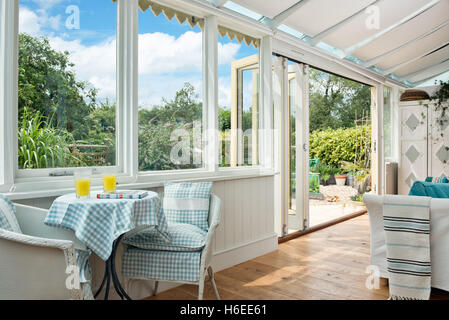 Ein Tisch zum Frühstück mit Gingham-Tischtuch, in einem großen Wintergarten mit Türen, die an einem sonnigen Tag zum Garten geöffnet sind. UK Stockfoto