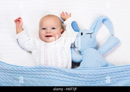 Lustige kleine Baby tragen eine warme Strickjacke, spielen mit Spielzeug Hase auf weißem Kabel stricken Decke in sonnigen Kinderstube. Stockfoto