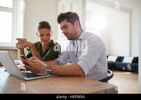 Aufnahme des jungen Paares mit Blick auf Handy Laptop zusammen am Tisch sitzen. Junger Mann und Frau mittels zu Hause smart phon Stockfoto