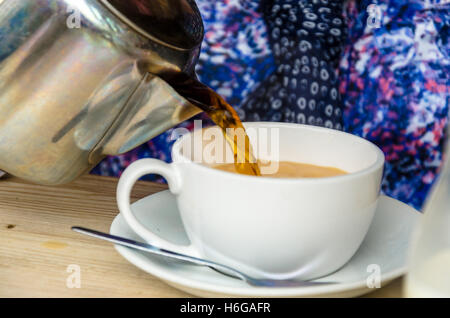 Eine Dame ergießt sich eine Tasse Tee aus einer Edelstahl-Teekanne in eine Tasse. Stockfoto