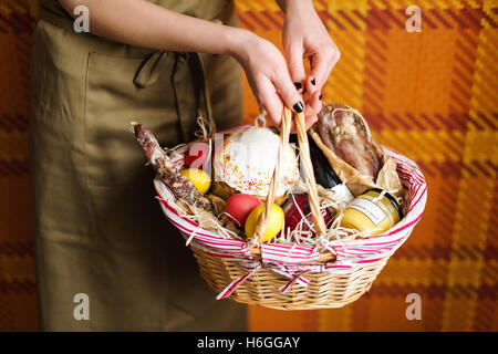 Weibliche Hände halten Korb mit bunten Eiern, Kuchen, Rotwein, Hamon oder ruckartig und trockenen geräucherte Wurst. Essen-Geschenk-set für das Osterfest. Stockfoto