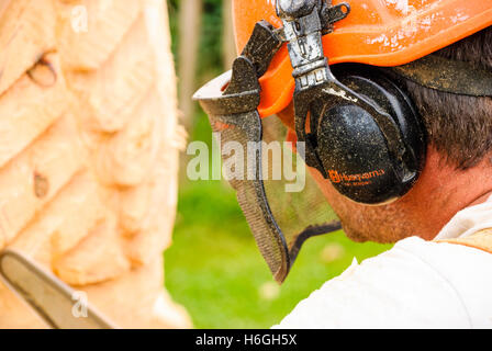 Ein Mann trägt einen Husqvarna Schutzhelm mit Gehörschutz und Gesichtsmaske, während er eine Kettensäge, eine große hölzerne Skulptur schnitzen verwendet. Stockfoto