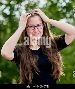 Junge Erwachsene Frau mit langen Haaren, Brille und lächelt beim erreichen Sie mit ihr Arme und Hände auf den Kopf Stockfoto