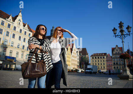 Zwei attraktive Freundinnen stehen auf gepflasterten Platz der schönen Stadt. Eine der Frauen in dunkle Brille, andere bedeckt ihre Augen mit Stockfoto