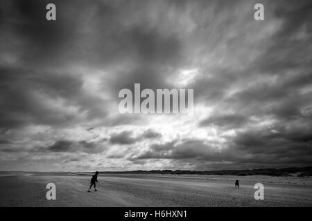 Frau und ihr Hund unter einem Gewitterhimmel an einem windigen Strand, Dornweiler an der North Norfolk-Küste. Stockfoto