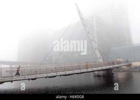 Die Menschen gehen über die Drehbrücke in Canary Wharf, London, wie Nebel oben auf den Gebäuden verdeckt. Stockfoto