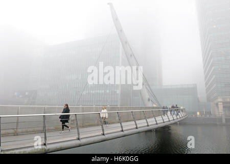 Die Menschen gehen über die Drehbrücke in Canary Wharf, London, wie Nebel oben auf den Gebäuden verdeckt. Stockfoto