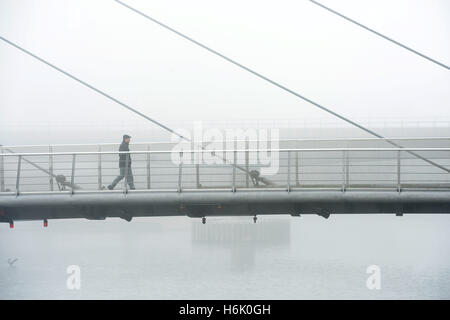 Die Menschen gehen über die Drehbrücke in Canary Wharf, London, wie Nebel oben auf den Gebäuden verdeckt. Stockfoto