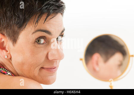 Nahaufnahme einer schwarzhaarigen Frau mittleren Alters mit runden Make-up-Spiegel in weißes Licht in die Kamera schaut. Stockfoto