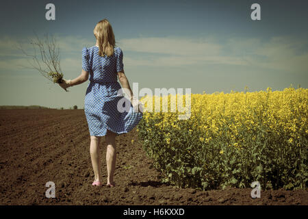 Rückansicht des kaukasischen blonde Frau mit blauen Polka Kleid hält ein Blumenstrauß steht auf Boden in einem Raps-Feld Stockfoto