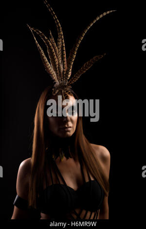 Gebräunte weibliche Modell im Amazonas Superhelden-Outfit mit Feder Kopfschmuck, dramatischer Schatten Stockfoto