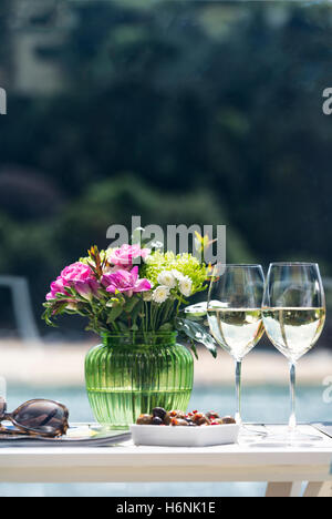 Zwei Gläser Weißwein mit Vase mit frischen Blumen und einer Schüssel mit Oliven auf der Seite von Pool an einem heißen Sommertag Stockfoto