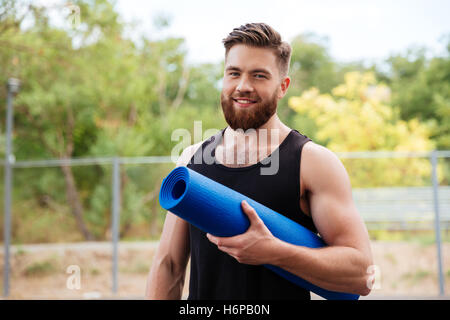 Handsome bärtiger junger Sportler halten Yogamatte und Blick auf Kamera im freien Stockfoto