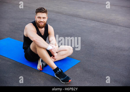 Lächelnde junge bärtigen Sport Mann sitzen und ruhen auf blau Fitness-Matte im freien Stockfoto