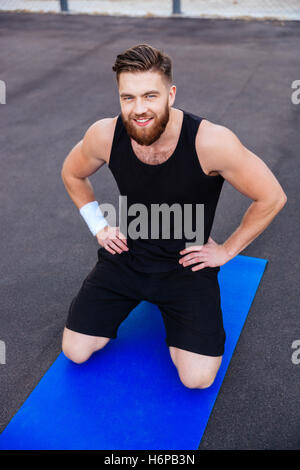 Glücklich lächelnd bärtiger Fitness Mann dabei auf blauen Matte im Freien Training Stockfoto