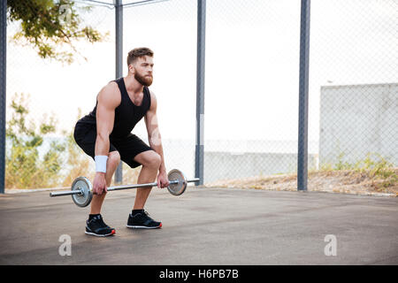 In voller Länge Portrait von ein ernster junge bärtige Mann Sportler trainieren und im freien Hantel heben Stockfoto