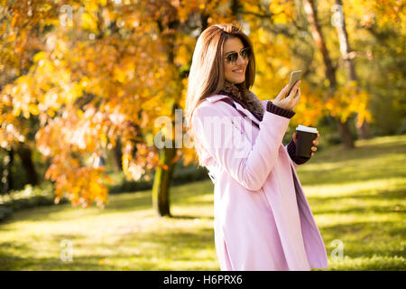 Junge Frau in den rosa Mantel im Gespräch über das Smartphone und hält Kaffee in der hand im Herbst Park gehen Stockfoto