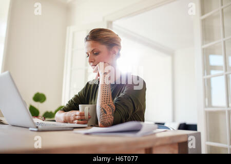 Innenaufnahme der jungen Frau mit Kaffee mit Blick auf Laptop. Frau sitzt am Tisch arbeiten. Stockfoto
