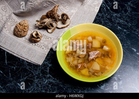 Pilzsuppe in grüne Platte auf einem schwarzen Stein Hintergrund. Einige getrocknete Steinpilze oder weiß Waldpilzen auf braunem Tuch. Stockfoto