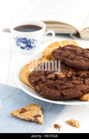 Große gemischte Schokoladenkekse auf einer Platte auf einem rustikalen Tisch mit schwarzem Kaffee und offenes Buch im Hintergrund. Stockfoto