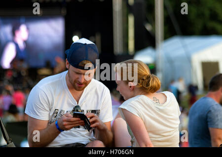 Junger Mann auf einem Musikfestival Überprüfung seiner Smartphone, beobachtet von einer jungen Frau Begleiter. Stockfoto