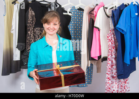 Fröhliches junges Mädchen steht mit einem roten großen Geschenk Hintergrund hängen Dinge auf Kleiderbügeln Stockfoto