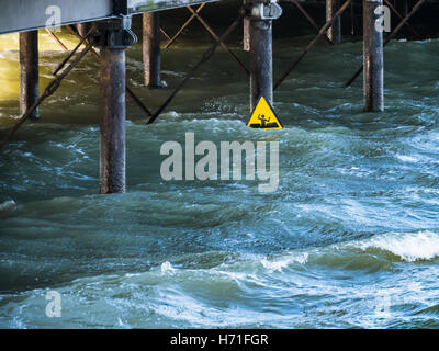 Ein gelbes Warnschild für weichen Schlamm sieht aus wie ein Mann winken für Hilfe vor dem Ertrinken unter einem Pier in tiefen kalten Wasser auf einem w suchen Stockfoto