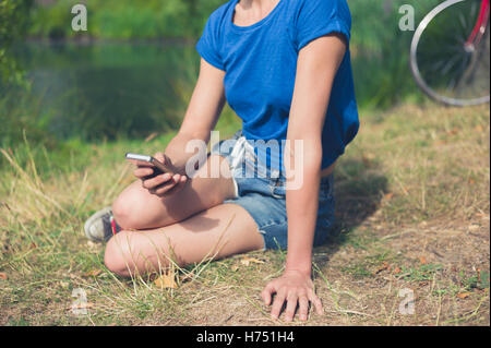 Eine junge Frau sitzt am Wasser in einem Park und ist ein Smartphone mit Stockfoto