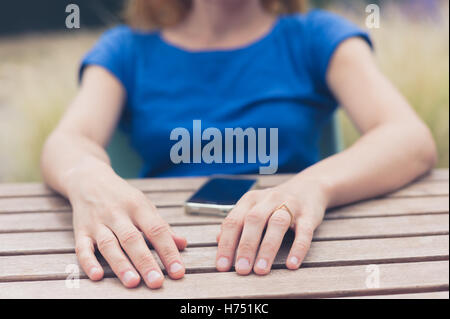 Eine junge Frau sitzt am Tisch im Freien und ist mit ihrem Smartphone an einem Sommertag Stockfoto