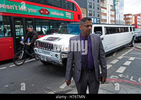 Asiatischer Mann auf Commercial Road im East End sieht auf der Straße als eine weiße Stretch-Limousine Hummer geht mit einer Gruppe von Menschen in London, England, Vereinigtes Königreich. Stockfoto