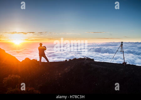 beleuchtete Silhouette Mann unter ein Foto eines Sonnenuntergangs über den Wolken aus einem Felsen mit hellen Sonne auf der linken Seite Stockfoto