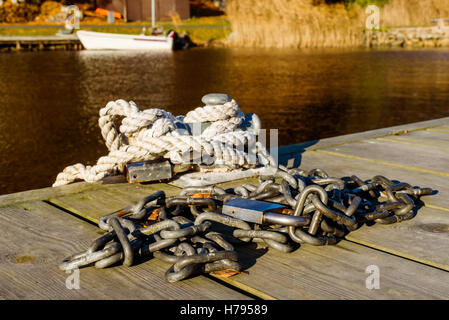 Stahlkette mit Vorhängeschloss auf hölzerne Pier. Seil und Vorhängeschloss auf Poller. Unscharfen Hintergrund mit festgemachten Boot und Küste. Stockfoto