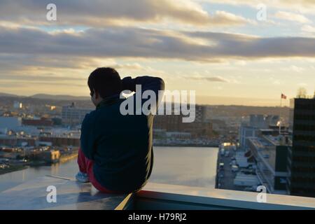 Ein Mann sitzt auf einem Felsvorsprung der einen Wolkenkratzer mit Blick auf die Stadt bei Sonnenuntergang. Stockfoto