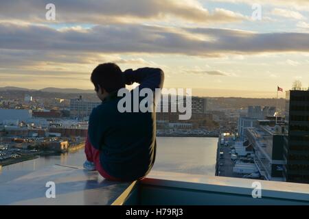 Ein Mann sitzt auf einem Felsvorsprung der einen Wolkenkratzer mit Blick auf die Stadt bei Sonnenuntergang. Stockfoto